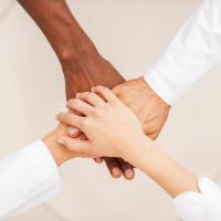 hands of different skin colours (white, olive, brown) clasping in a circle with a white tiled background.