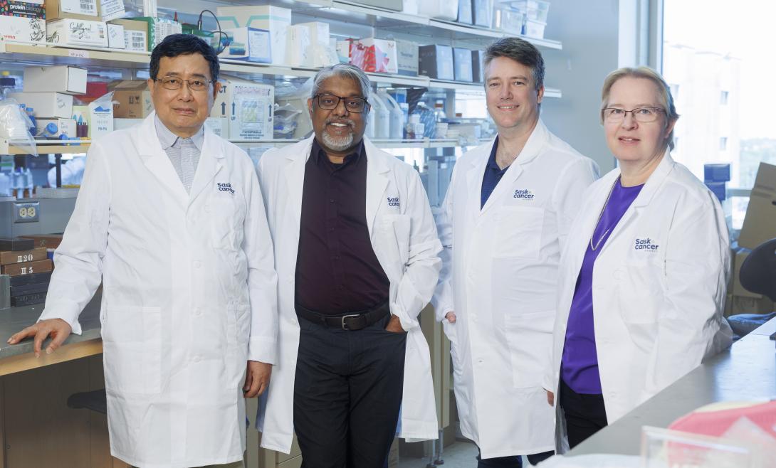 4 individuals left to right (3 men and 1 woman) wearing white lab overcoats standing in front of laboratory lab equipment.